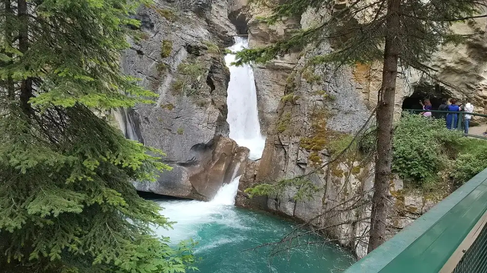A waterfall dropping into a green pool in Johnston Canyon with a walkway and visitors on the rock ledge beside it