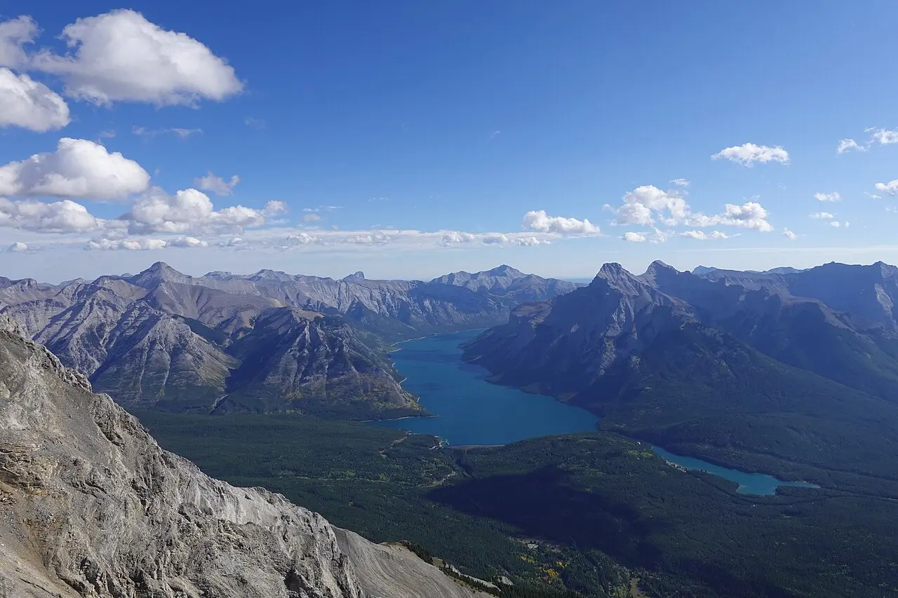 Lake Minnewanka stretching between forested mountain ridges, seen from a high ridge with clouds above