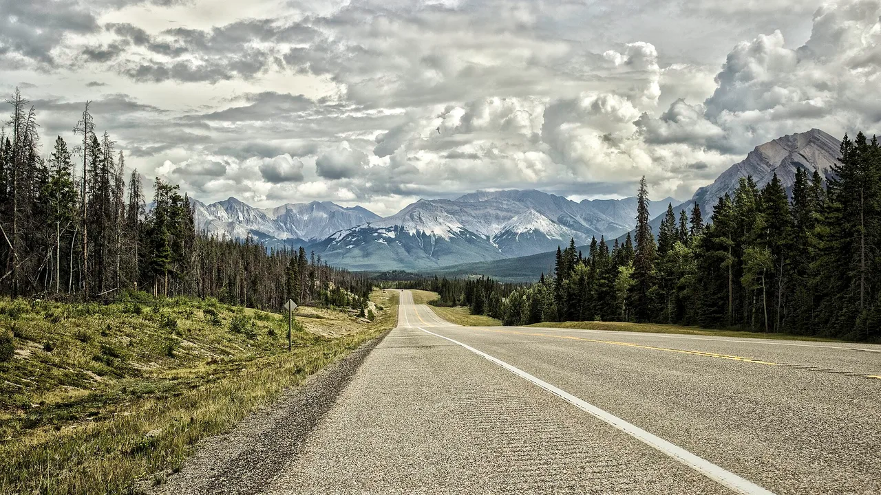 An empty two-lane highway curving through the Canadian Rocky Mountains under a dramatic cloudy sky