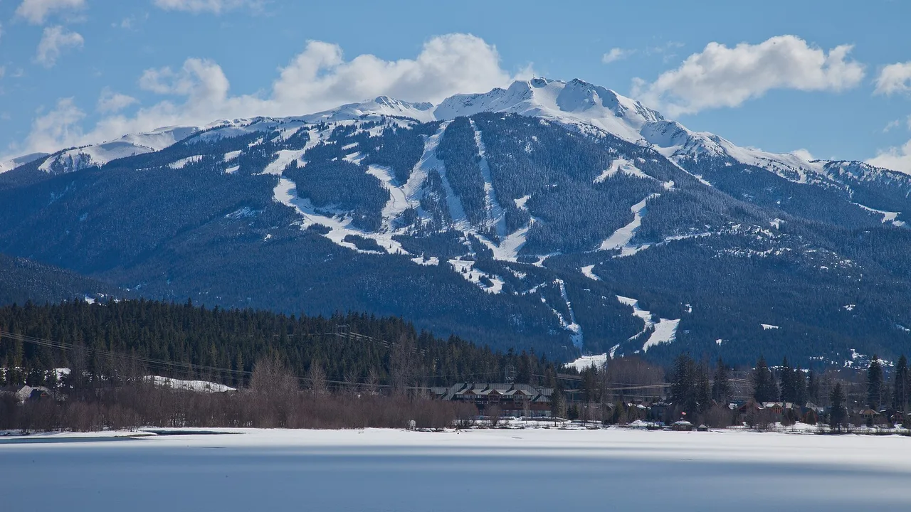 Whistler Mountain and its ski runs above a partly frozen Green Lake under a blue sky with clouds in British Columbia