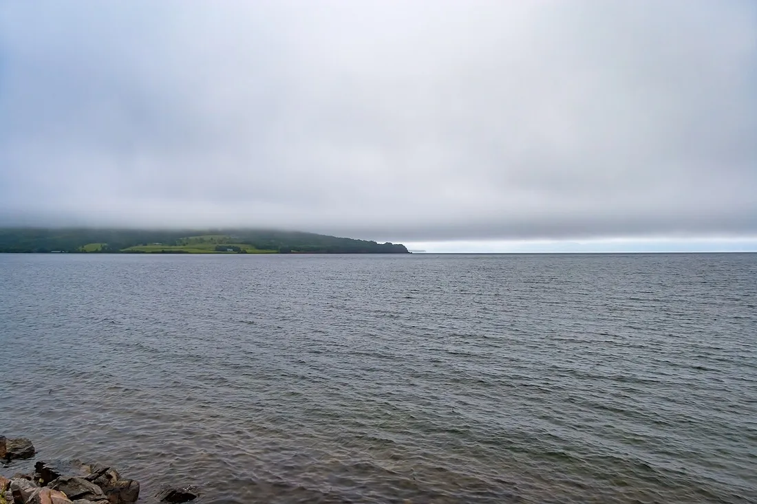 The calm Bras d'Or Lake at Baddeck under low grey cloud, with a farm on a distant green point