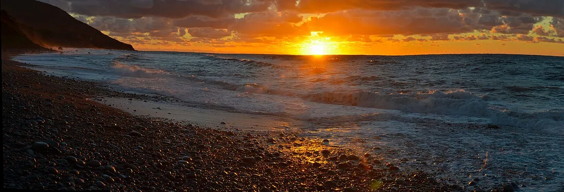 A fiery orange sunrise over a pebble beach and breaking waves below dark highlands near Cape North