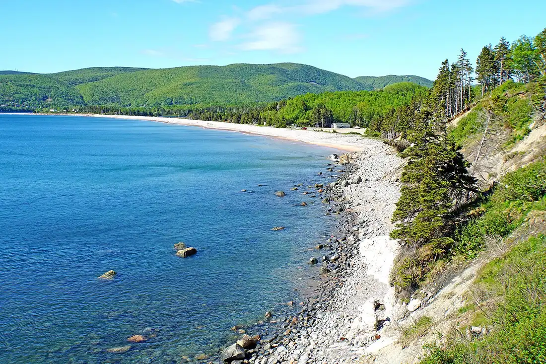 A curving white-pebble beach and clear turquoise water backed by forested highlands at Ingonish