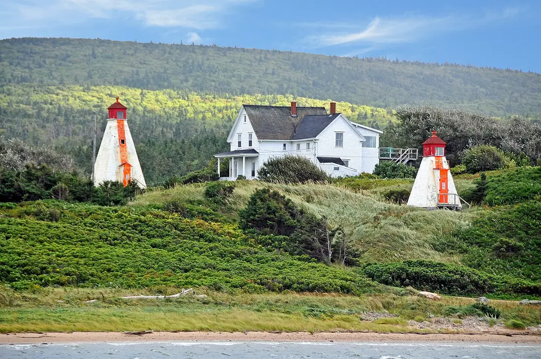 Two red-and-white range light towers beside a white house on a grassy bluff above the beach at Margaree Harbour