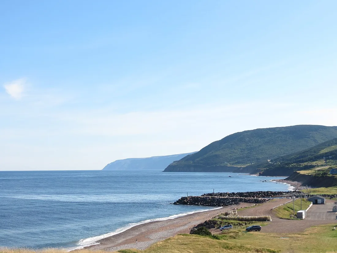 A grey-pebble beach and breakwater below green highlands under a clear sky at Pleasant Bay