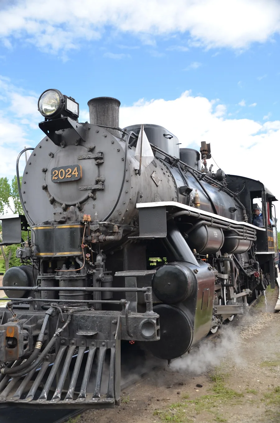 A black steam locomotive numbered 2024 standing under steam on the tracks at Heritage Park with a driver leaning from the cab