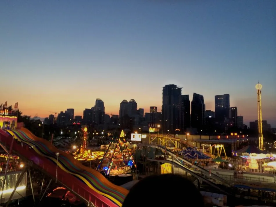 The Calgary Stampede midway lit up at dusk with rides in the foreground and the downtown skyline on the horizon