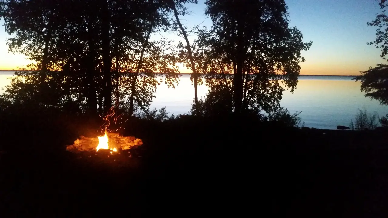 A tent and camp chairs pitched on a low bluff above a calm lake at dusk in Manitoba