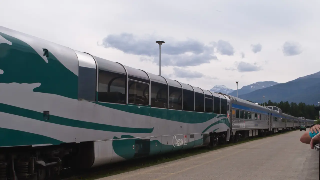 A VIA Rail train with a dome car standing at Jasper station with mountains behind