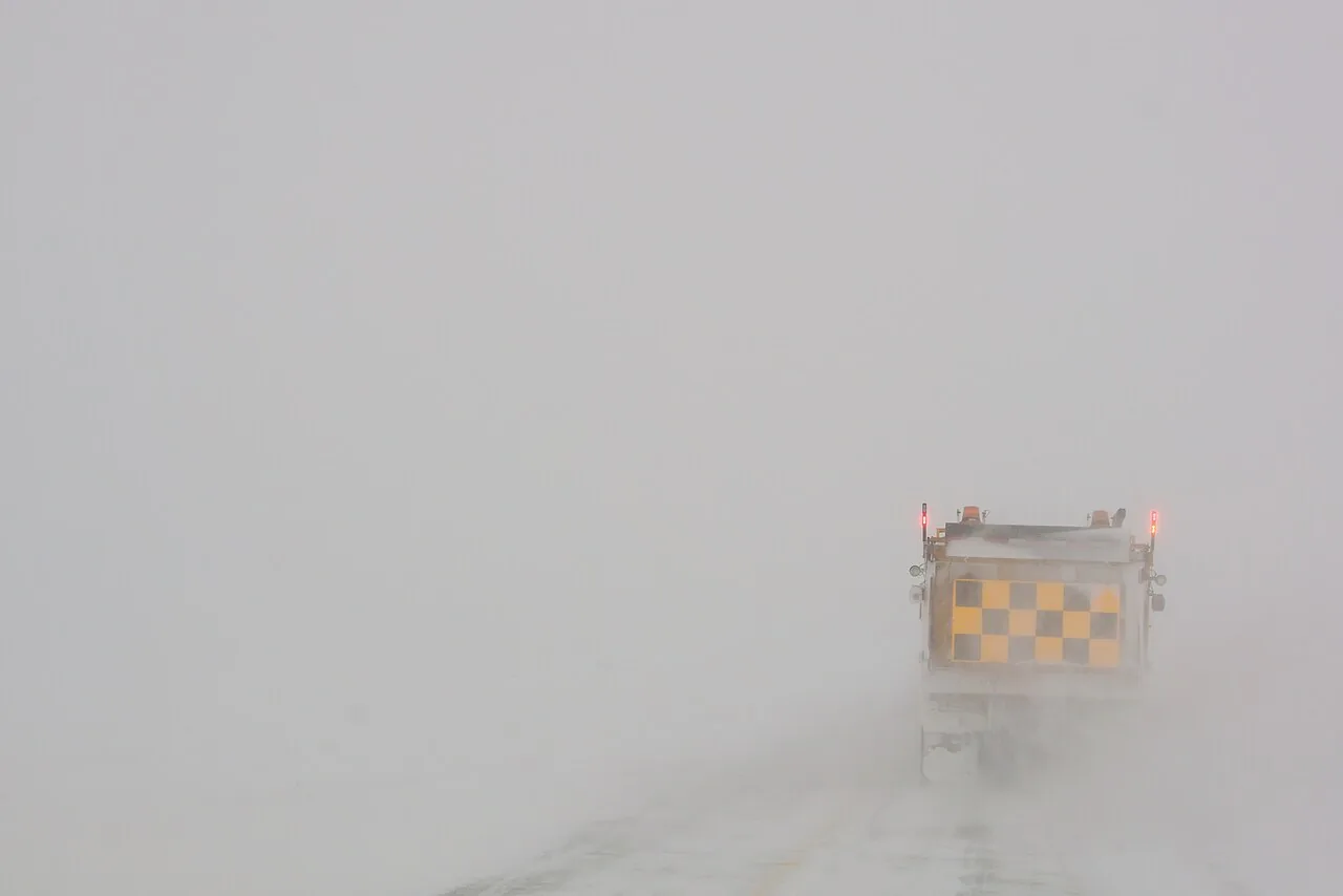 A snowplow truck barely visible ahead in a highway whiteout with blowing snow