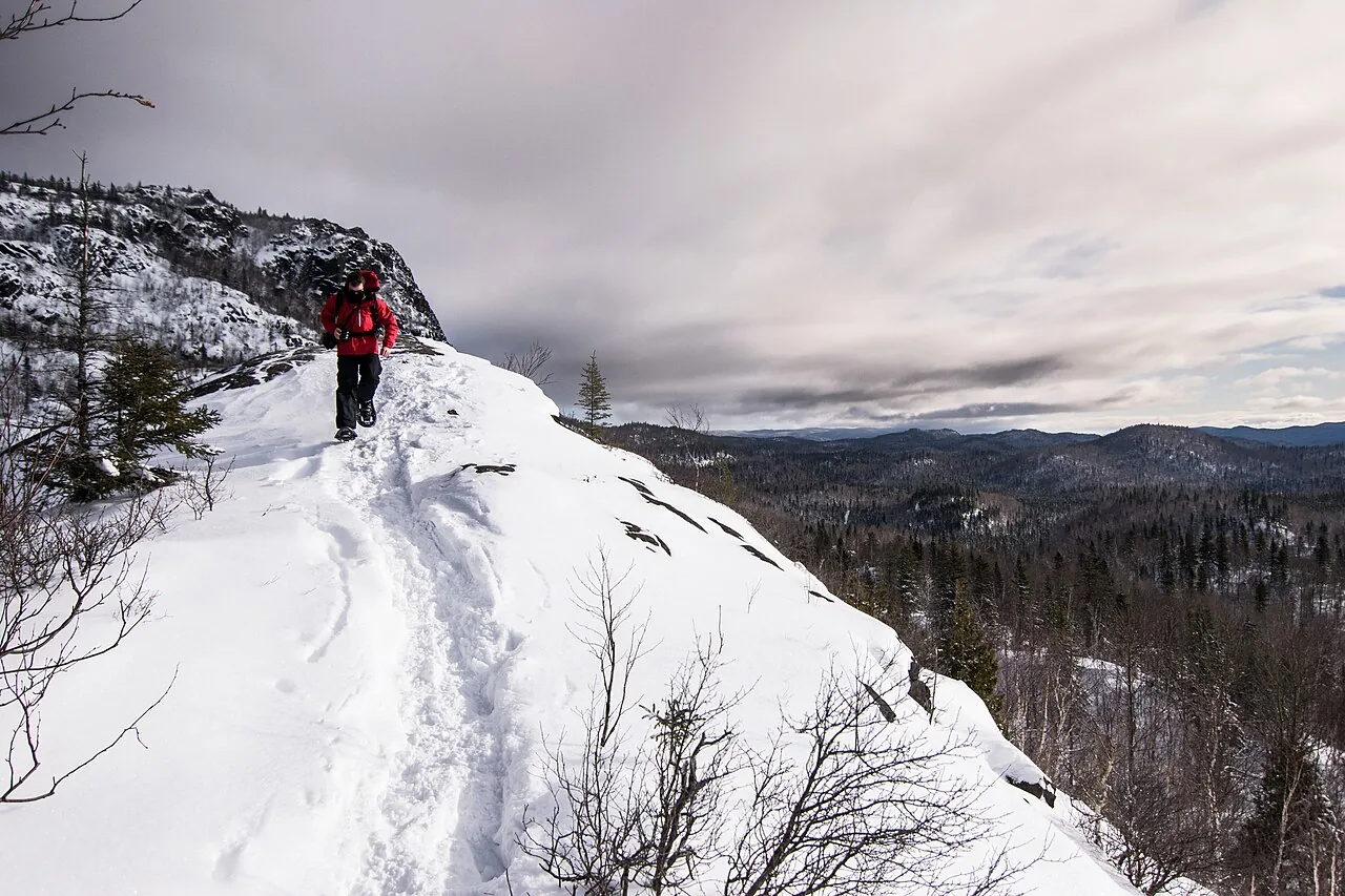 A snowshoer in a red jacket on a snowbound ridge in the Monts-Valin, Quebec, with forested hills below
