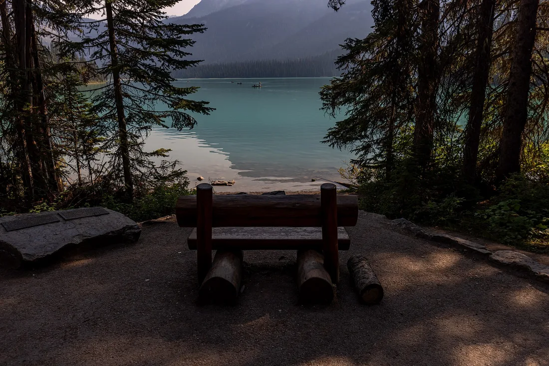 The green water of Emerald Lake in Yoho National Park with a wooden bench on the shore and forested peaks behind