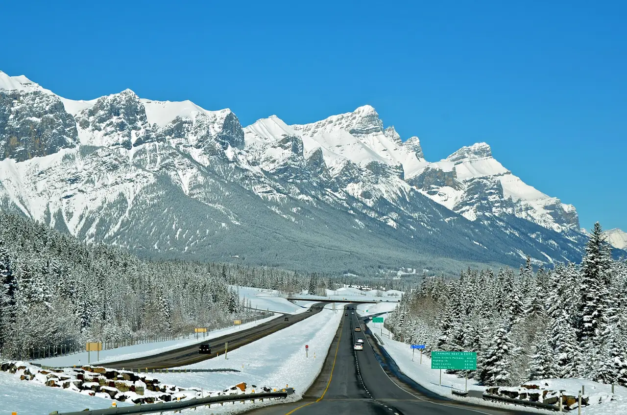 Highway 1 heading west toward Canmore with clear pavement, snowy forest on both sides, a snow-covered ridge behind and a green sign counting down the Canmore exits