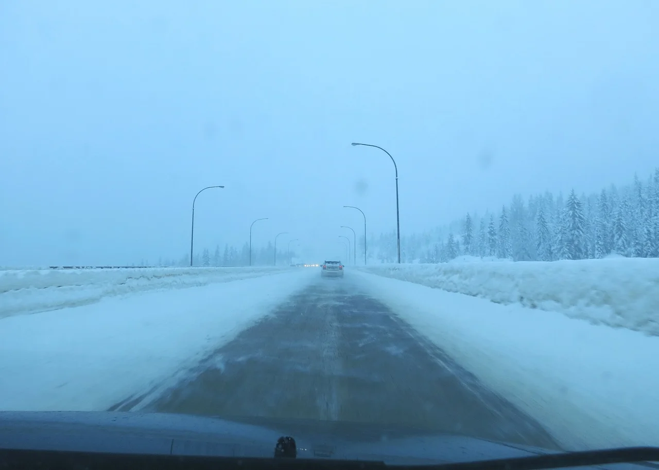 A snow-banked highway in heavy snowfall seen through a windshield, with one car ahead and streetlights lining the road
