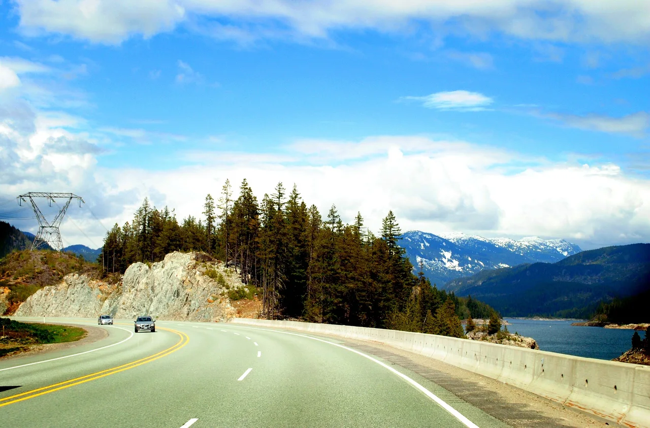 Highway 99 curving along a mountain lake with snow-dusted peaks in the distance and two cars on the road