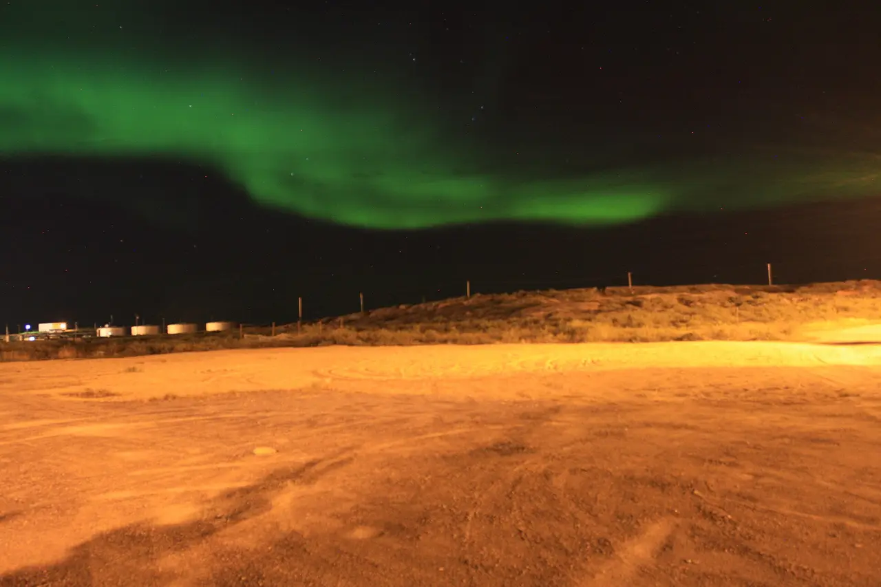 A green aurora arc over open ground and fuel tanks on the edge of Churchill, Manitoba, at night
