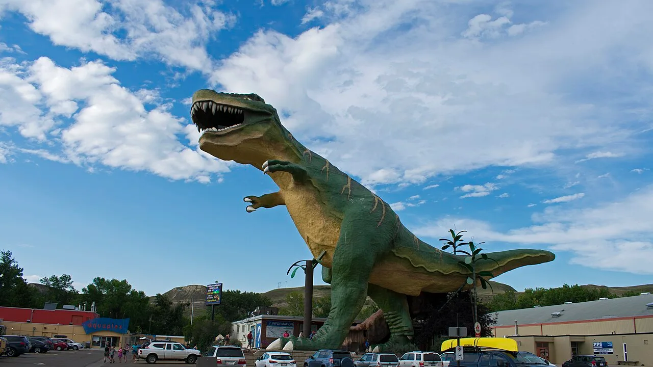 The giant World's Largest Dinosaur statue towering over a parking lot in downtown Drumheller, Alberta, with badlands hills behind it