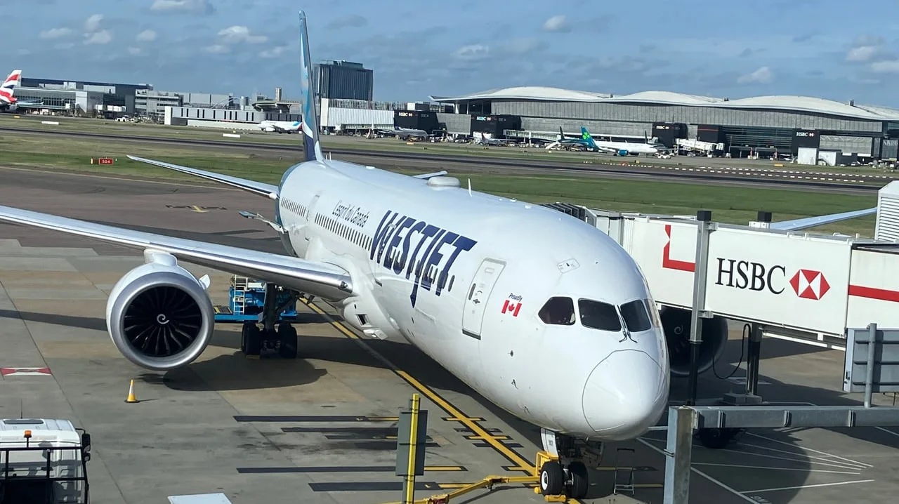 A WestJet Boeing 787-9 Dreamliner parked at a jet bridge at London Heathrow, with the terminal and taxiways behind