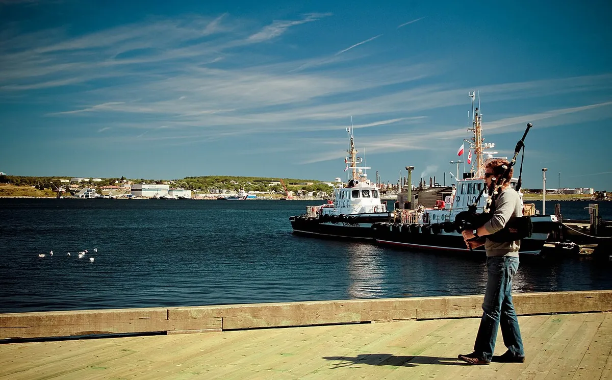 A bagpiper in jeans playing on the wooden boardwalk of the Halifax waterfront with pilot boats and the harbour behind