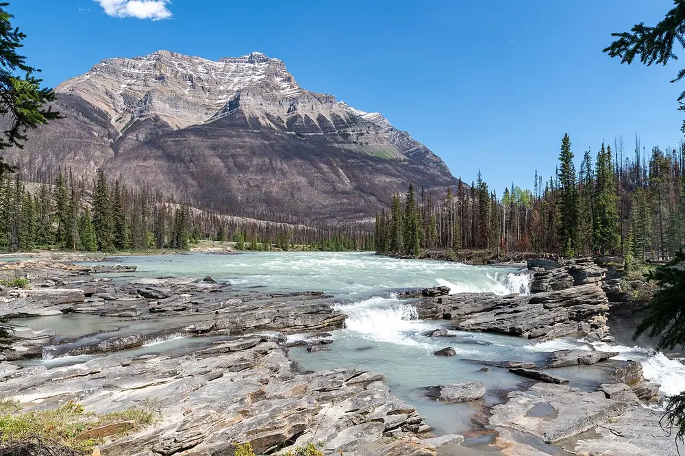 The Athabasca River pouring over layered rock shelves below a steep dark mountain face
