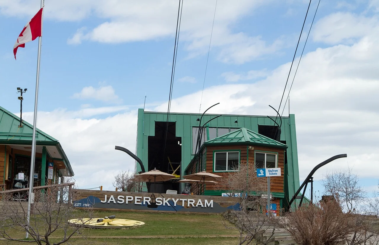 The green Jasper SkyTram lower station building with its cables rising out of the roof, a Canadian flag and a sign pointing to the ticket window