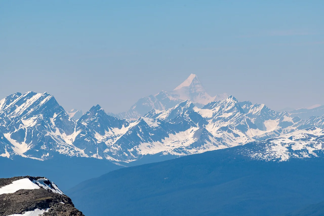 Mount Robson rising as a pale pyramid behind rows of snowy peaks, seen in haze from the top of The Whistlers