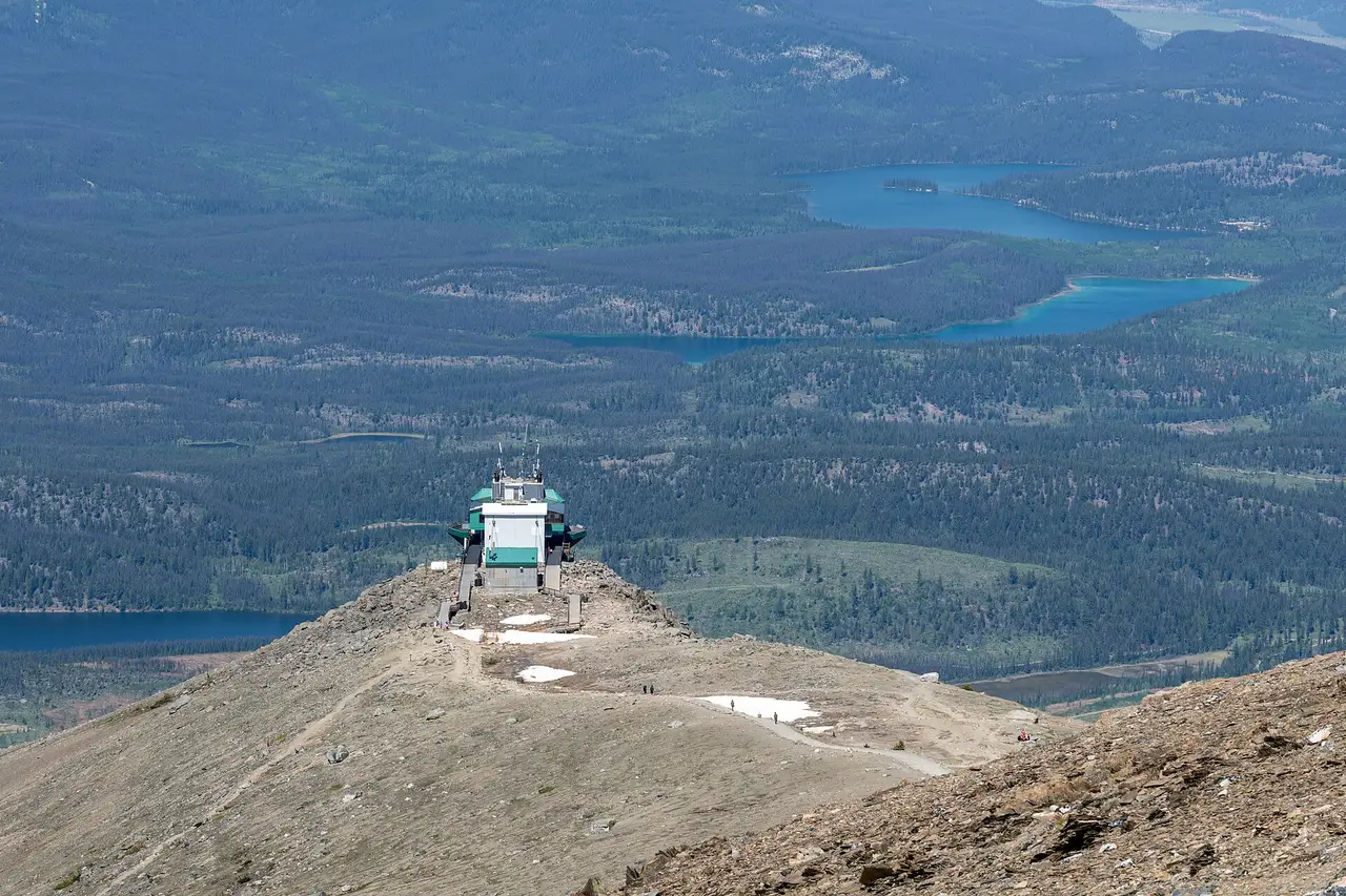 The SkyTram upper station on a bare ridge seen from higher up the summit trail, with walkers on the path and two blue lakes in the forest far below