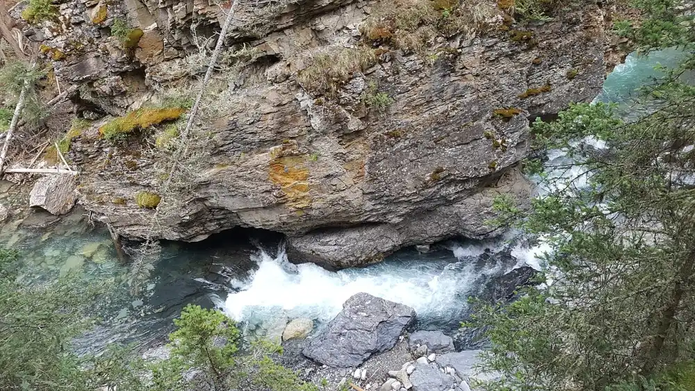 Turquoise creek water rushing beneath an overhanging limestone wall in Johnston Canyon