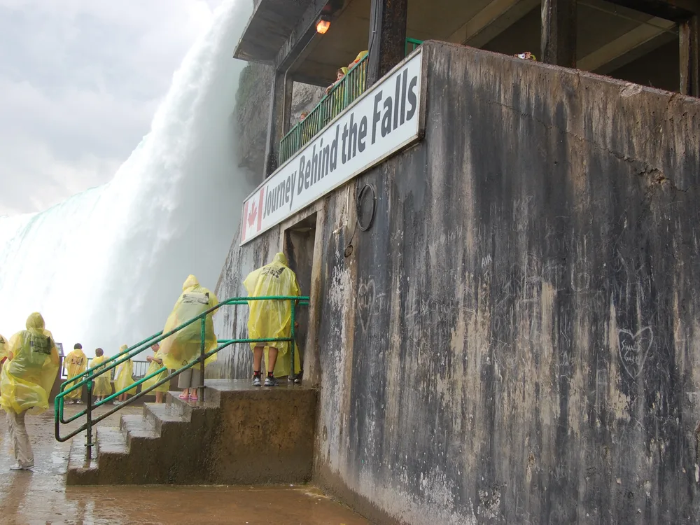 The Journey Behind the Falls sign on the concrete building at the foot of the Horseshoe Falls, with visitors in yellow ponchos on the steps