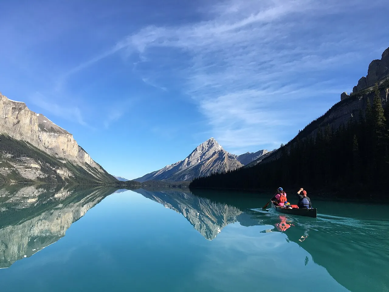 A family paddling a rented canoe on the still, mirror-like water of a Rockies lake with a glacier-topped peak reflected ahead