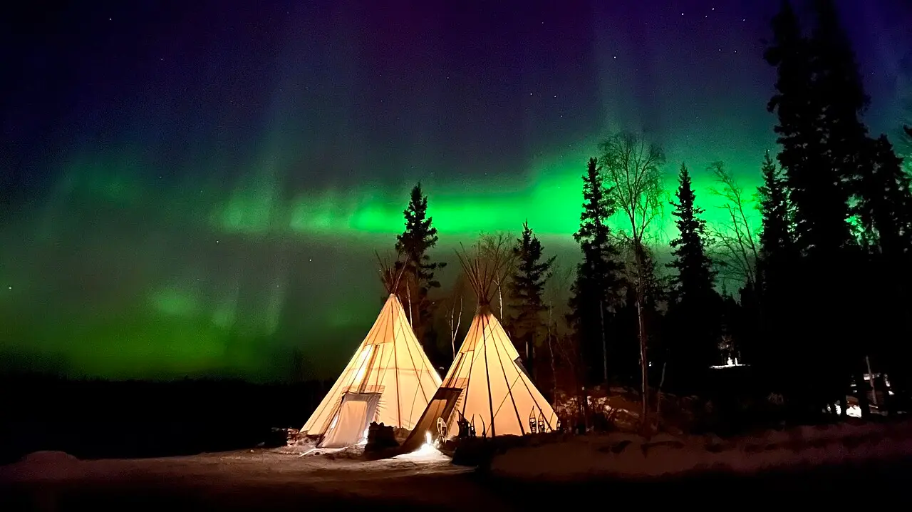 Teepees silhouetted under a green aurora arcing across the night sky at Cassidy Point near Yellowknife