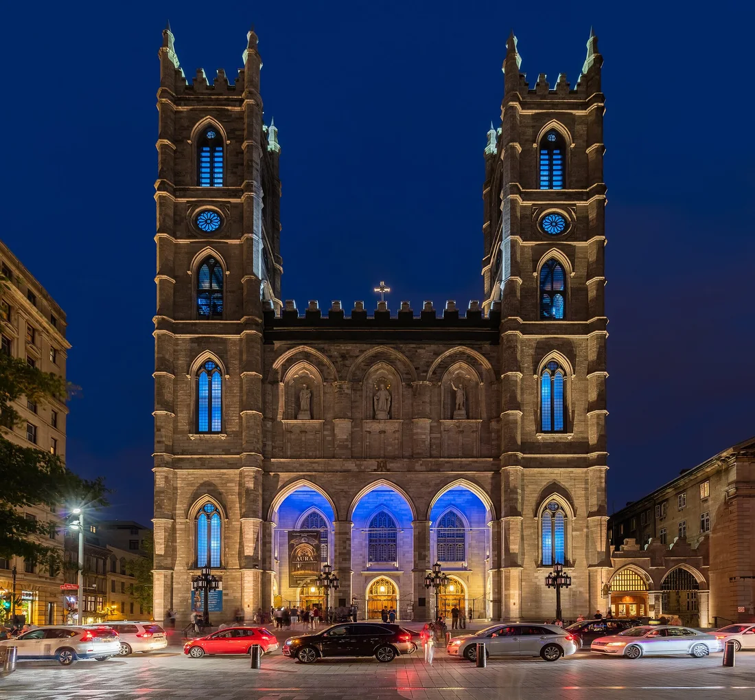 The twin-towered facade of Notre-Dame Basilica after dark with blue light in the three entrance arches and an AURA poster at the door