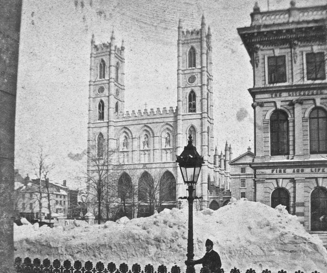 Black-and-white 1870s photograph of the twin-towered Notre-Dame church seen across Place d'Armes, with a snowbank and a gas lamp in the foreground