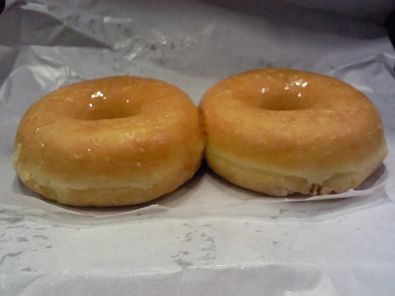 Two classic glazed doughnuts on wax paper, the simple style Lee's Donuts has hand-rolled since 1979