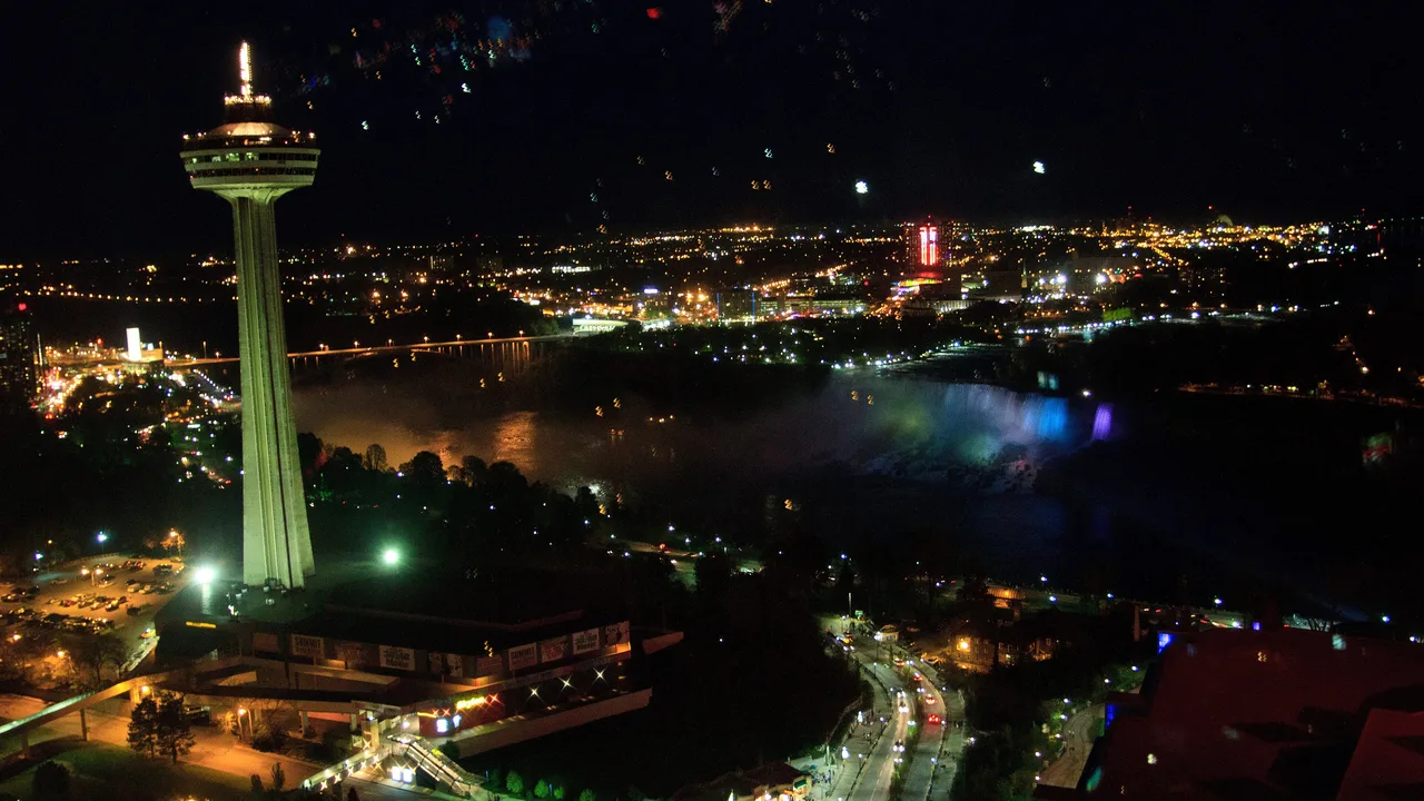 The floodlit Skylon Tower at night, with the illuminated American Falls, the Rainbow Bridge and the lights of Niagara Falls behind it