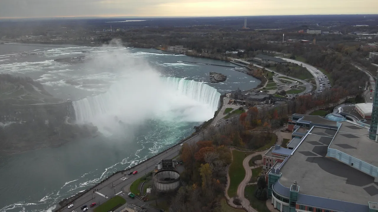 The Horseshoe Falls seen from high above, a plume of mist rising from the curve, with Table Rock, the Niagara Parkway and autumn trees along the Canadian shore