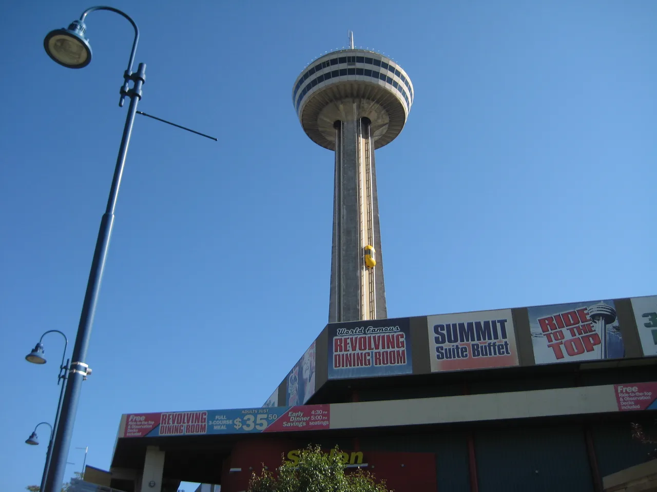 The Skylon Tower rising above its entrance building, with banners for the Revolving Dining Room, the Summit Suite Buffet and the Ride to the Top, and a yellow elevator halfway up the shaft