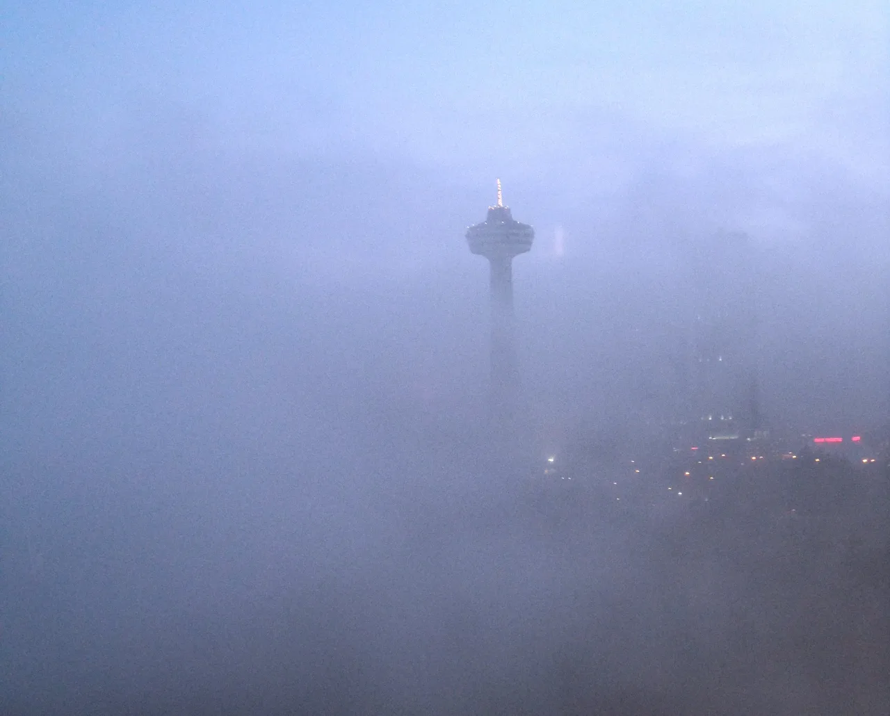 The top of the Skylon Tower barely showing through thick blue-grey fog, with a few street lights glowing below