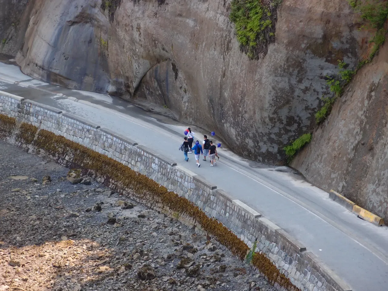 The paved seawall path running below a rock face in Stanley Park, with a tidal algae line on the stone and walkers heading around the point