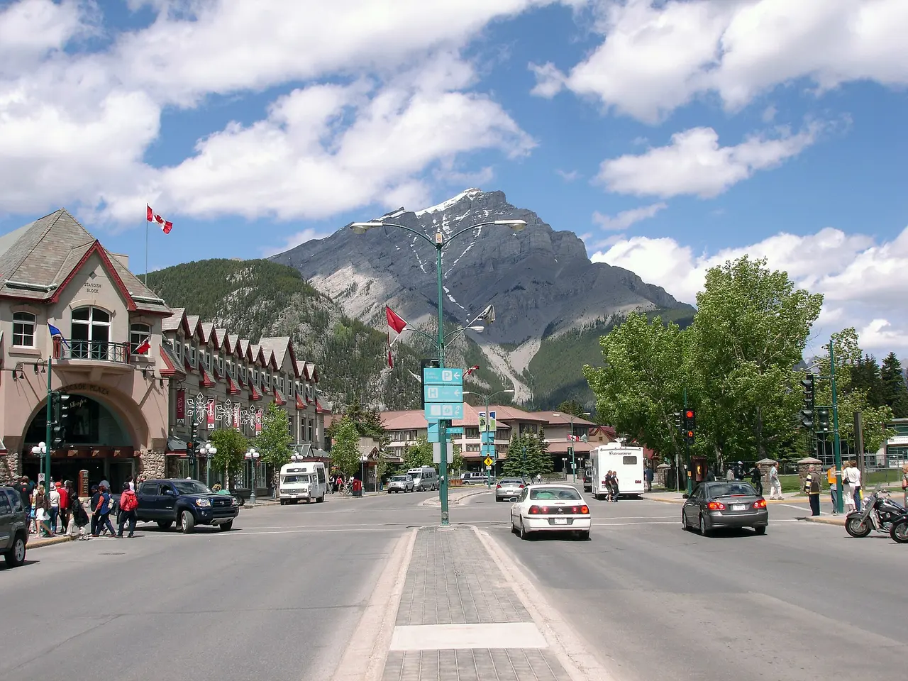 Banff Avenue looking north towards Cascade Mountain, with shops, flags, cars and pedestrians on a summer day