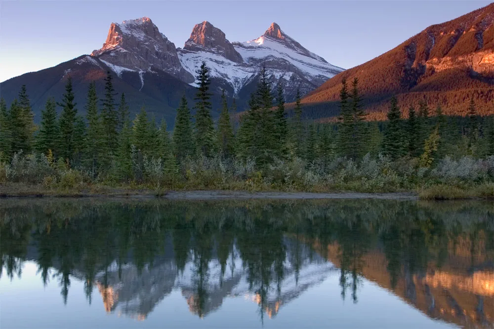 The three peaks of the Three Sisters reflected in the still Bow River at dawn just east of Canmore
