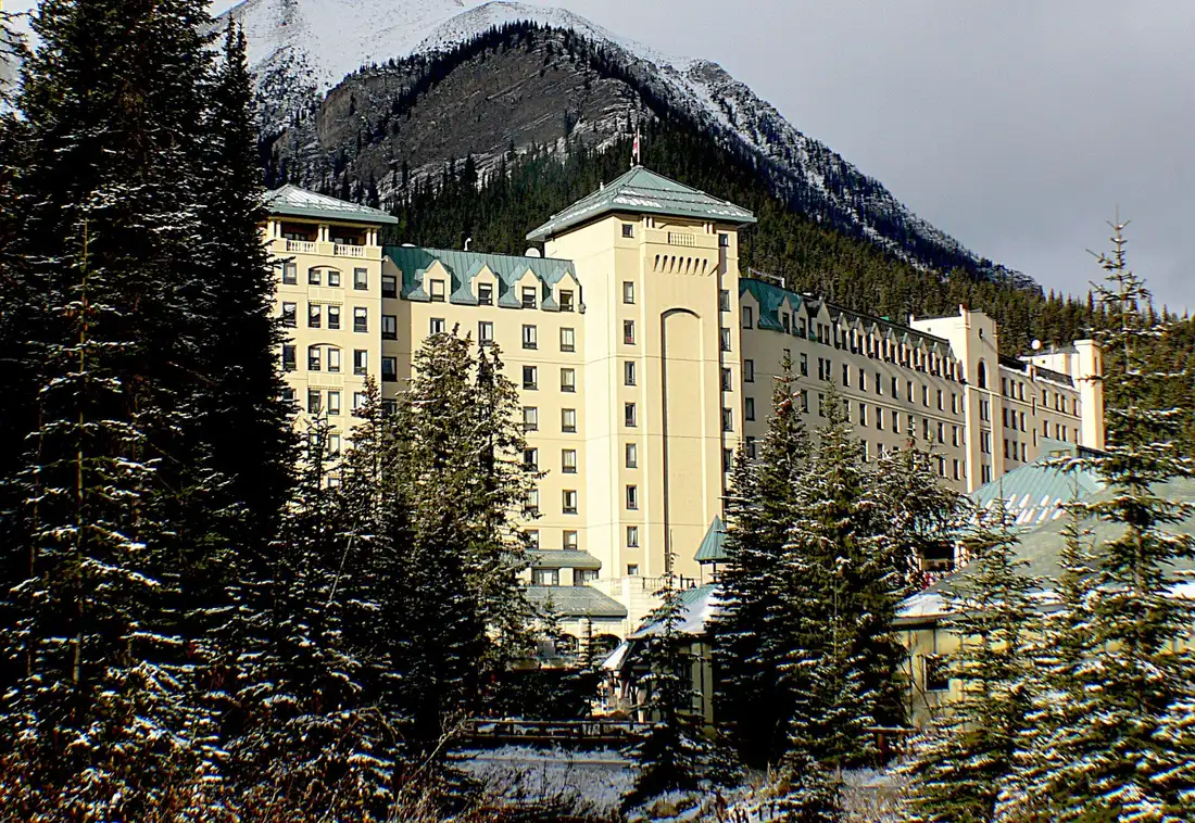 The pale stone bulk of the Fairmont Chateau Lake Louise seen through snow-dusted spruce trees below a dark mountain ridge