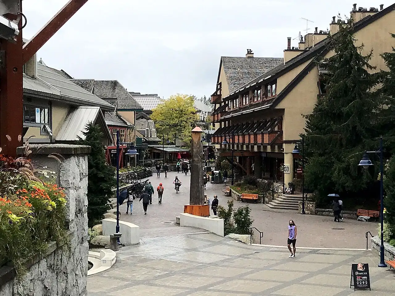 Pedestrians walking through the Village Common in Whistler past shops and a carved wooden totem